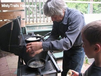 A man is cooking Halibut in a pan on a grill and there is also a boiling pan of water on the grill as well. A boy is watching the man cook.