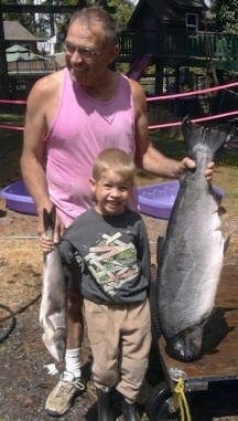 A man in a pink muscle shirt is standing behind a boy. The man is looking to the left and he is holding a Salmon in his left hand. The boy is holding up a fish in his right hand.