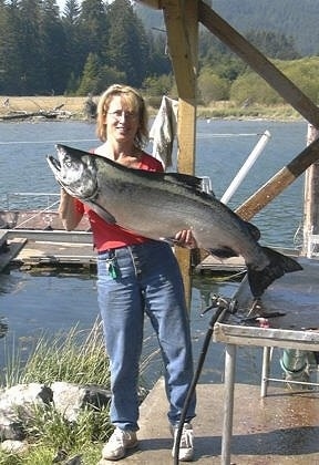 A lady in glasses and a red shirt is standing on a stone platform near a body of water and she is holding up a large Salmon fish.
