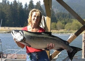 A lady in glasses and a red shirt is holding up a large Salmon in her hands.