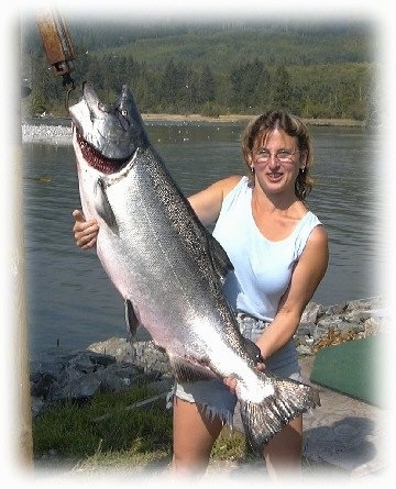 A lady in glasses is holding up a Salmon with mechanical aide. The fish is huge, almost as large as the lady.