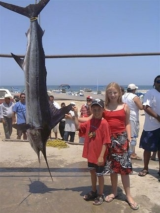 A boy in a hat and a blonde haired girl are standing next to a Striped Marlin that is hanging upside down from a line. They are both touching the large back fin of the marlin fish. The fish is larger than the kids.
