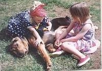 A girl in a blue polka dot dress and a girl in a pink polka dot dress are laying around a black and tan Bloodhound dog sleeping in grass.