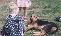 A child in a blue and white polkadot dress is kneeling in front of a black and tan Bloodhound that is laying on its right side. There is another child in a pink polka dot dress behind the dog.