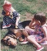 A girl in a blue polka dot dress is putting a Stethoscope on the side of a Bloodhound that is laying in grass. There is a girl in a pink polka dot dress sitting next to the Bloodhound.