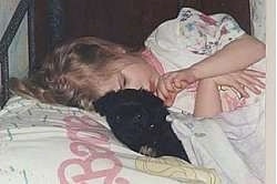 Close up - A girl is sleeping in a bed with her head on a Barbie pillow and next to her is a Havanese puppy looking forward.