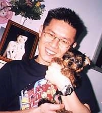 A man is standing next to a framed picture of a lady and he is holding a tiny black and tan Silky Terrier puppy in his arms.