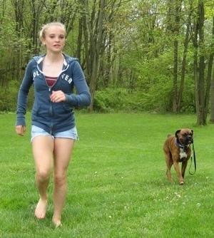 Amie running and Bruno the Boxer is following behind her with his leash in his mouth