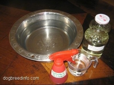 A Silver bowl with clear liquid in it, a red spray bottle, a measuring cup filled with liquid and a glass jar are sitting on a wooden table