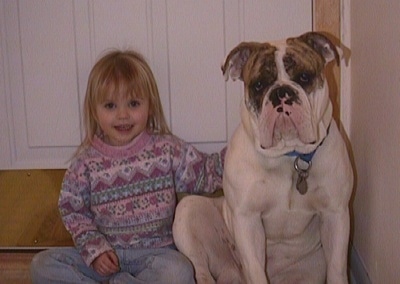 A white with brown and black Bulldog is sitting next to a little girl on the floor leaning against a door.