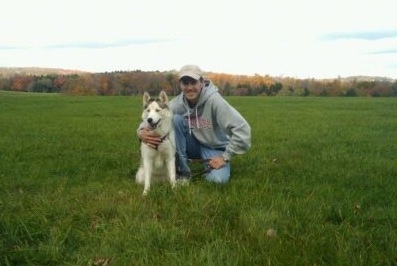 A black, grey and white Siberian Husky is sitting in a field next to a man in a grey hoodie who has its arm around the side of the dog.