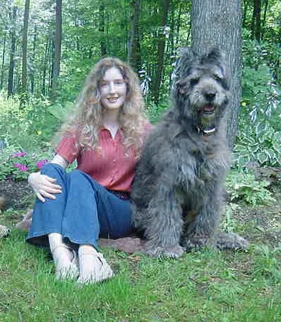 Dixie the Bouvier des Flandres sitting in the grass next to a lady with flowers and trees in the background