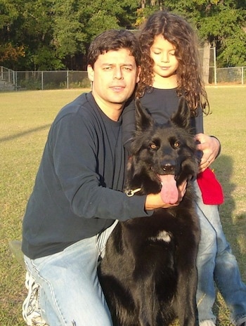 A black German Shepherd is sitting outside in the outfield of a baseball field in front of a man and a little girl who are both wearing blue shirts.