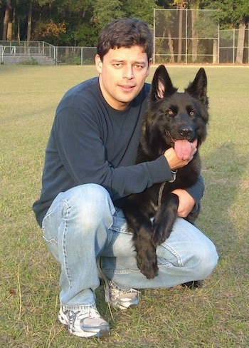 A man in a blue shirt has his arms wrapped around the body of a black German Shepherd in the outfield of a baseball field.