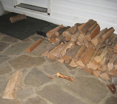 A stacked pile of split logs on a stone porch with a few logs separated from the pile and a dog bone laying in front of it in front of a white farm house.