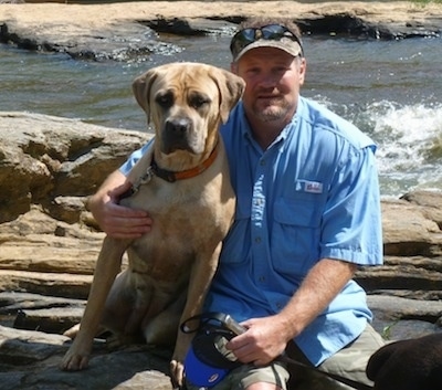 Cassie the Cane Corso Italiano is sitting next to her owner on a rock and there is a body of water behind them