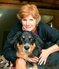 A lady with short red hair is standing behind a black, tan and white Australian Shepherd/Rottweiler/Border Terrier mix. Her chin is on the back of the dogs head and her arms are over the side of the dog.