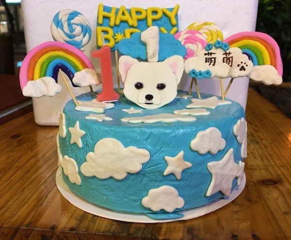 A round light blue birthday cake with white clouds and stars on it. There are rainbows, lollipops, a white dog head, the number one and a Happy Birthday decoration on top of it.