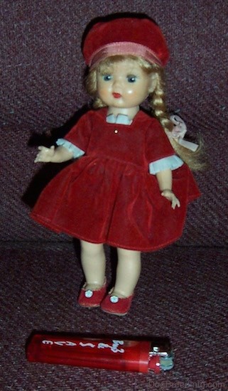 A small doll with long blonde braids wearing a red velvet hat and dress and red shoes standing up with a red lighter in front of her for size comparison.