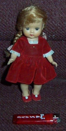 A small doll with long blonde braids wearing a red velvet dress and red shoes with small white flowers on them standing up with a red lighter in front of her for size comparison. The doll has blue eyes, a small nose and red lips.
