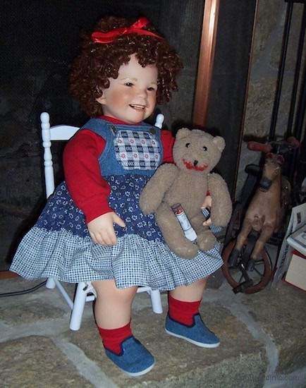 A realistic looking doll of a little girl with short curly ringlet curls in her hair sitting in a chair