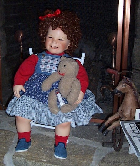 A doll with Shirley Temple curly hair with a red ribbon on her head with a teddy bear on her lap sitting in a white chair.