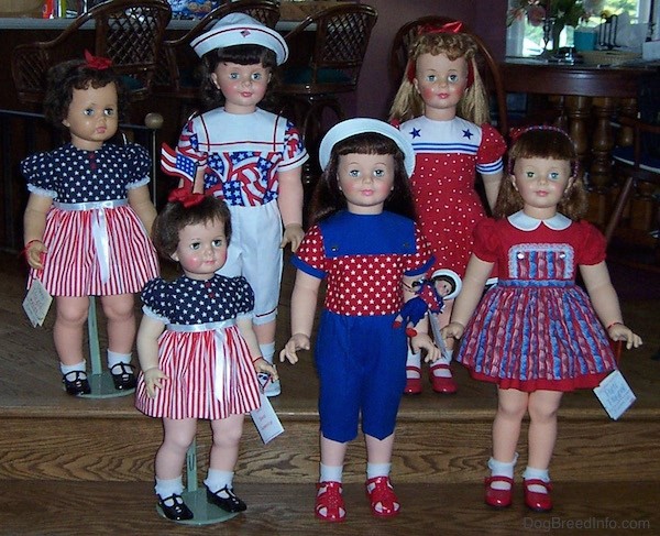 Front view - Six realistic looking large dolls dressed in red, white and blue standing on wooden steps. Two dolls have white hats and two dolls have red ribbons and two have red hair bands.