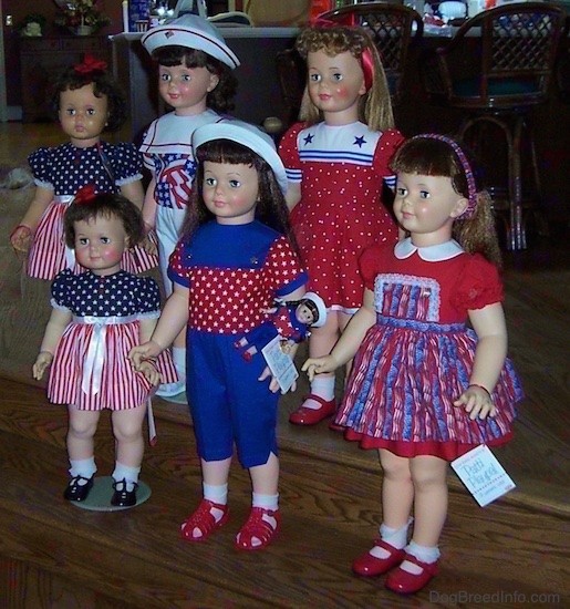 Side view - Six realistic looking large dolls dressed in red, white and blue standing on wooden steps. Three dolls have red shoes, two have black shoes and one has on white shoes. One larger doll wearing a white hat has a small identical toy doll attached to her shirt.