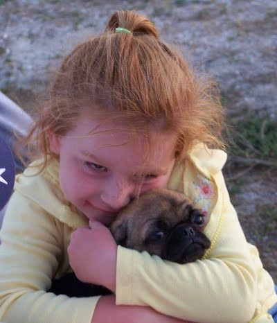 A little girl with red hair and a yellow jacket hugging a tan and black puppy