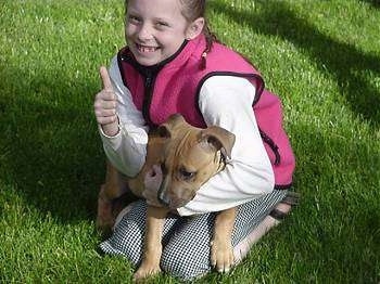 A girl is kneeling in grass wearing a hot pink vest and a white shirt and she has a tan Pit Bull puppy laying across her lap. She is giving a thumbs up, the puppy is looking to the left.