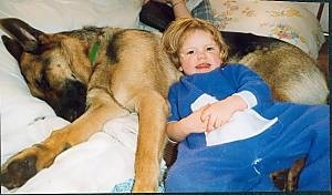 A black with tan German Shepherd dog is sleeping on its side next to a child in a blue onesie who is laying on his back with his head on the dog's belly.
