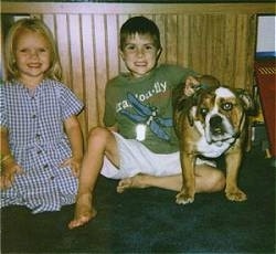 A boy and a girl are sitting on a dark green carpet in front of a small wooden wall. Next to the boy is a brown and white Bulldog. They all are looking forward.