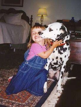 A girl in a pink shirt and blue jean suspenders is on her knees hugging a Dalmatian