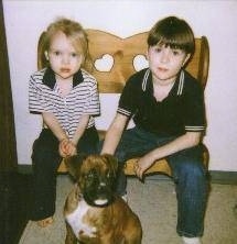 A boy and a girl are sitting next to each other on a wooden bench. There is a brown with white and black Boxer puppy sitting on a carpet in front of them. The Boxer is looking to the right and the children are looking forward.