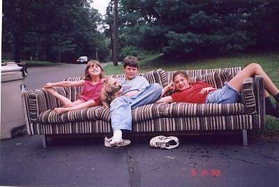 A boy and a tan Cairn Terrier are surrounded by two girls on a couch. The couch is outside in the street. The Cairn Terrier dog is looking to the right.