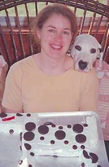 A Dalmatian has its head on the shoulder of a lady in a yellow shirt. There is a Dalmatian cake on a table in front of them