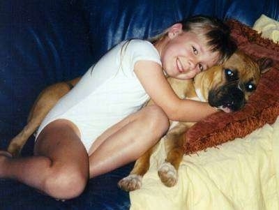 A smiling girl in a white onesie is laying on a couch on top of a brown with white Boxer.