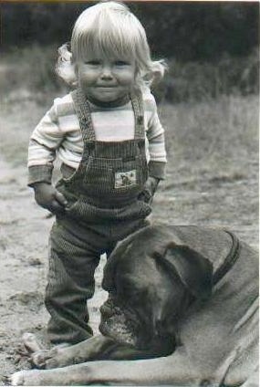 A black and white photo of a blonde toddler standing next to a Boxer that is laying down outside