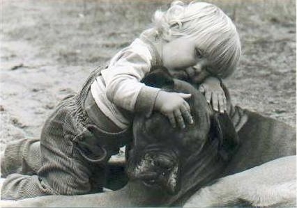 A black and white photo of a blonde toddler laying across the head of a Boxer dog outside in the dirt.