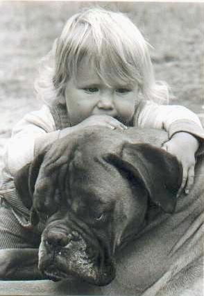 Close up - A black and white photo of a blonde toddler laying across the head of a Boxer dog outside in the dirt.