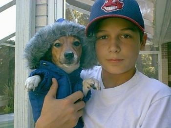 Front view - A white with tan Parson Russell Terrier dog is wearing a blue parka with a fuzzy hood and there is a boy wearing a blue and red Cleveland Indians baseball cap holding the dog up near his head.