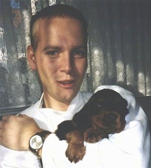 A black and tan Gordon Setter puppy is being held in the arm of a teenage boy wearing a white shirt and a watch.
