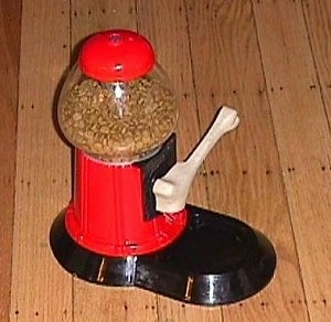 A Yuppy Puppy Treat Machine that looks like a red and black gumball machine with a handle shaped like a white dog bone sitting on a hardwood floor and it has dog food in it.