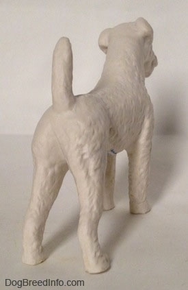 The back right side of a white bisque porcelain Airedale Terrier dog figurine that is unglazed. The dogs bak legs are straight and the tail is up high in the air.