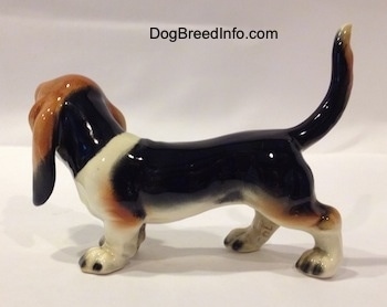 The left side of a black with white and brown Basset Hound figurine. The tail of the figurine is arched up.