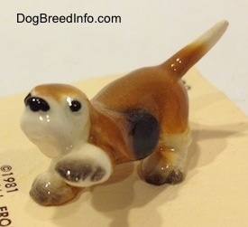 The front left side of a tan with black and white ceramic Basset Hound figurine that has its front left paw in the air. The eyes of the figurine are black circles.