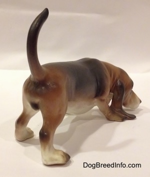 The back right side view of a brown and black with white porcelain Basset Hound figurine that is sniffing.