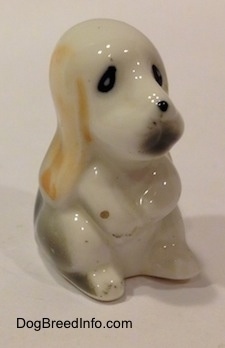 The front right side of a white with tan and black bone chine Basset Hound puppy figurine that is sitting on its hind legs. The figurine has poor details.