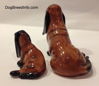 The back of two, a mother and a puppy, brown and black ceramic Basset Hounds figurines. The tails of the figurines are long.
