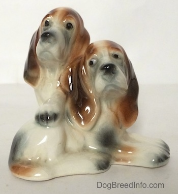A ceramic Basset Hound figurine that is two Basset Hounds, one laying across a surface and the other standing up across its back.